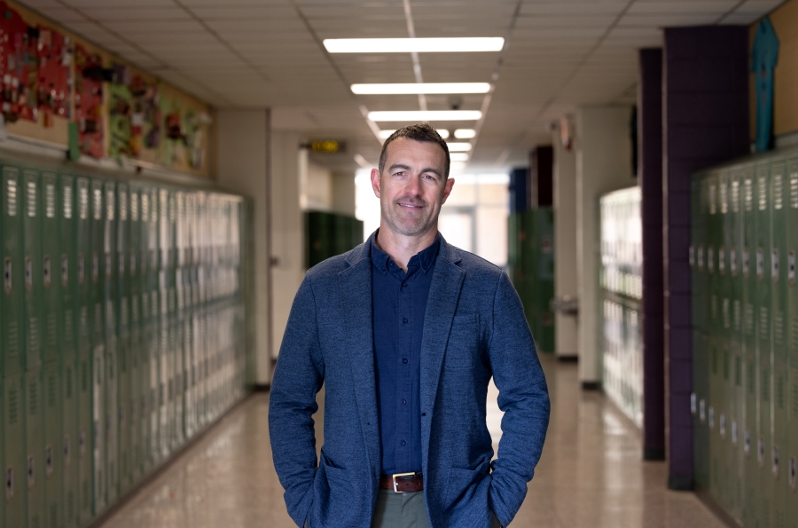 Photo of educator Caleb Coon - a man standing in a hallway full of lockers.